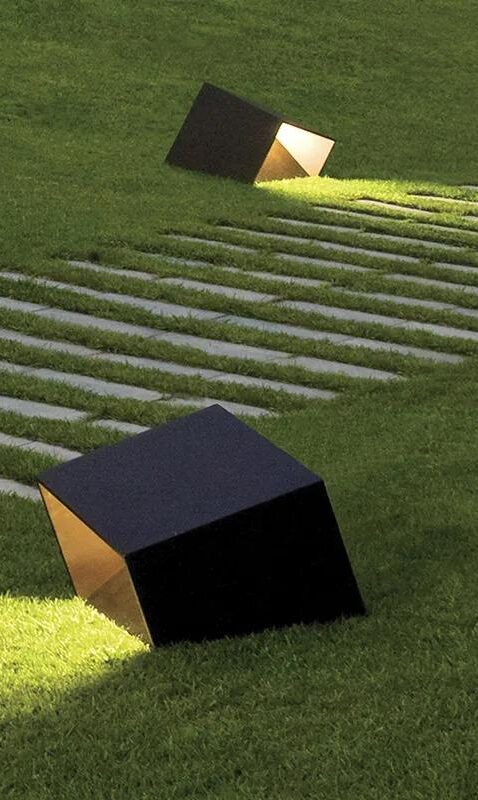 Zwei schwarze würfelförmige Außenleuchten von Burelbach, die teilweise in Gras eingebettet sind, beleuchten den Boden neben einem Steinweg aus rechteckigen Platten. Die Szene scheint in einem modernen Landschaftsgarten zu spielen.
