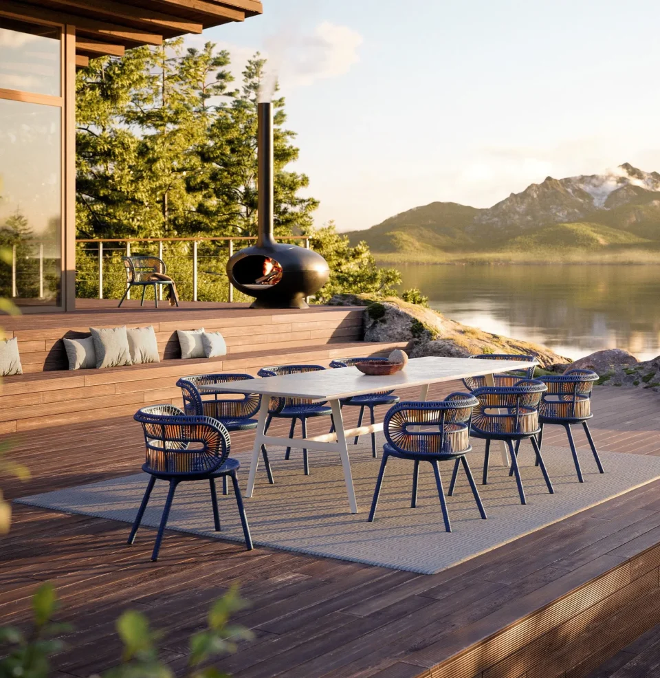 Ein moderner Essbereich im Freien von Gardendesign Burelbach mit blauen Stühlen und einem weißen Tisch auf einer Holzterrasse mit Blick auf einen See und die Berge, mit einer runden schwarzen Feuerstelle, Lounge-Kissen und üppigen Bäumen im Sonnenuntergangslicht.