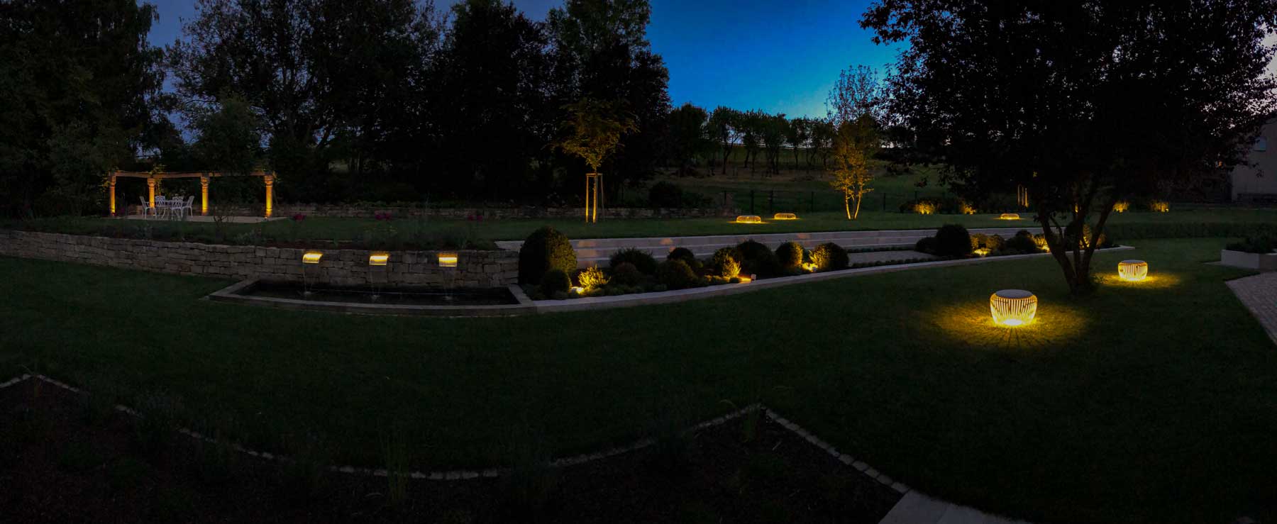 Ein Landschaftsgarten in der Abenddämmerung mit beleuchteten Bänken und Wegeleuchten, Steinmauern aus Burelbacher Handwerk, beschnittenen Sträuchern und einer Pergola im Hintergrund - inmitten von Bäumen und offenen Grasflächen werden Stile aus Luxemburg und Deutschland gezeigt.