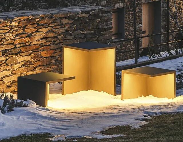 Drei moderne, würfelförmige Bänke von Burelbach leuchten warm mit Innenbeleuchtung in einem verschneiten Außenbereich und verbinden innovatives Gardendesign an einer Steinmauer und einem Gebäude mit Fenstern in Luxemburg und Deutschland.