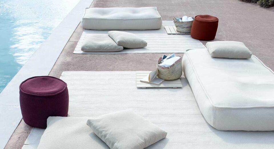 Sitzbereich im Freien am Pool mit weißen Bodenkissen, Gartenmöbeln, geflochtenen Körben, beigen Teppichen und einem runden kastanienbraunen Hocker auf Beton. Dieser ruhige und einladende Raum zeigt Gardendesign inspiriert von Deutschland und Luxemburg.