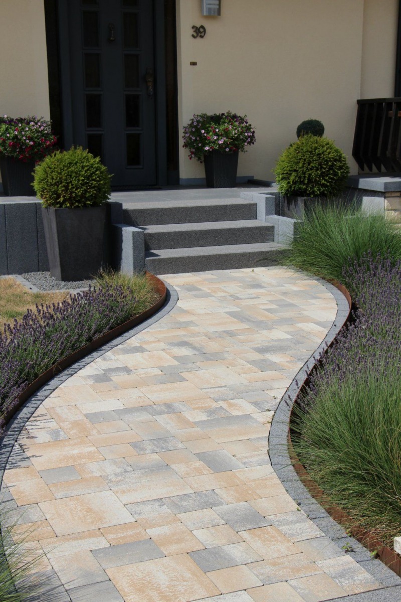 Ein geschwungener Steinweg führt zu einer Haustür mit Stufen, flankiert von grünen Sträuchern, Topfpflanzen und ordentlich geschnittenen Büschen - eine einladende Präsentation von Gardendesign und Burelbach Handwerk in einer sauberen, gepflegten Landschaft.