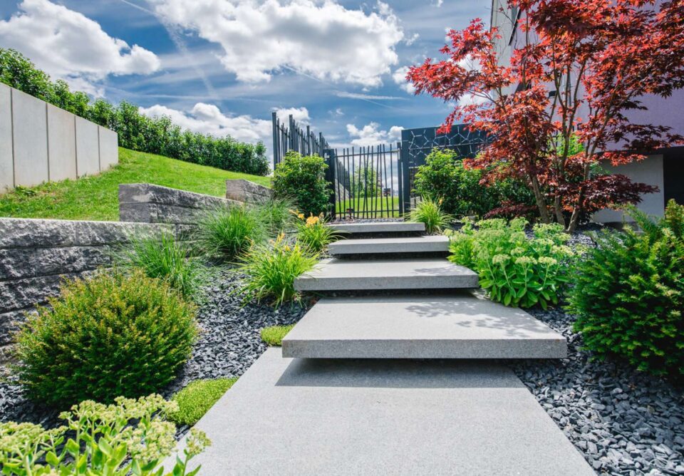 Ein moderner Gartenweg mit großen, flachen, handwerklich gefertigten Steinstufen führt durch üppige Grünpflanzen und Sträucher zu einem schwarzen Metalltor. Kräftiges rotes und grünes Laub rahmt den Weg unter einem teilweise bewölkten Himmel ein.