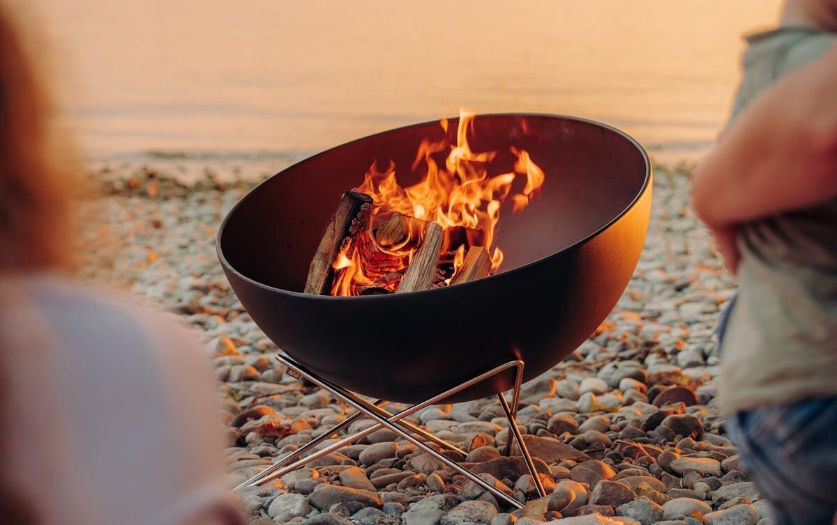 Eine Feuerstelle mit brennenden Holzscheiten sitzt bei Sonnenuntergang an einem felsigen Strand, zwei Personen genießen die Aussicht und das ruhige Wasser im Hintergrund - eine inspirierende Szene für die Gartengestaltung oder elegante Gartenmöbel-Arrangements.