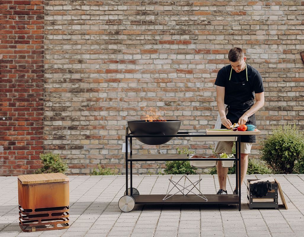 Ein Mann in einer Schürze präsentiert sein Handwerk, während er an einer Grillstation im Freien vor einer Backsteinmauer Speisen zubereitet, mit frischen Zutaten und Kochutensilien auf dem Tisch. Ein Holzhocker und üppige Gartenmöbel sorgen für Charme im Grünen.