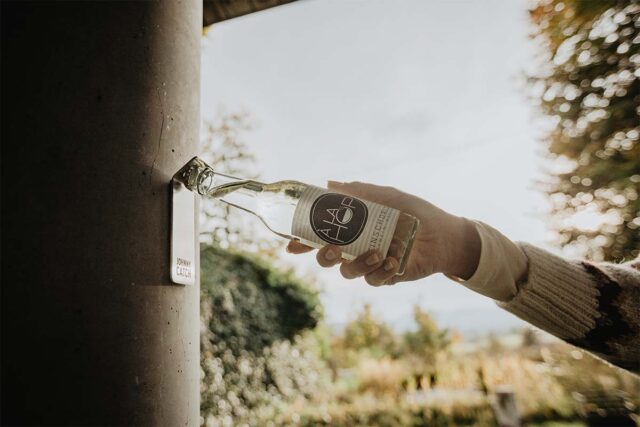 Eine Hand benutzt einen eingebauten Flaschenöffner an einem Betonpfosten, um eine Glasflasche mit der Aufschrift "A&P" zu öffnen. Die Szene, perfekt für Gartenmöbel-Liebhaber in Deutschland und Luxemburg, zeigt Grünflächen und Sonnenlicht im Hintergrund.