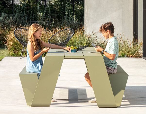 Zwei Kinder sitzen sich an einem modernen Burelbach Gartenmöbel-Tisch mit angeschlossenen Bänken gegenüber, essen und spielen ein Brettspiel. Das hohe Gras und die schwarzen Terrassenstühle vervollständigen die charmante Gartengestaltung im Hintergrund.