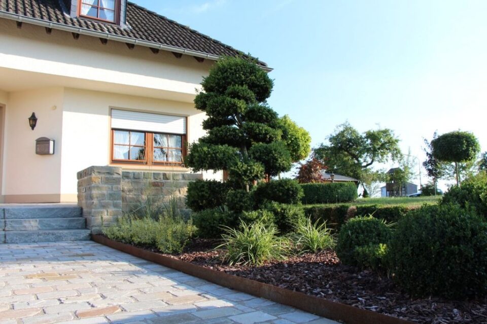 Ein ordentlich angelegter Garten vor einem Haus mit Burelbach Gartenmöbeln, einem beschnittenen Zierbaum, Mulch, grünen Sträuchern, Steinstufen und einer Kopfsteinpflaster-Auffahrt an einem sonnigen Tag.
