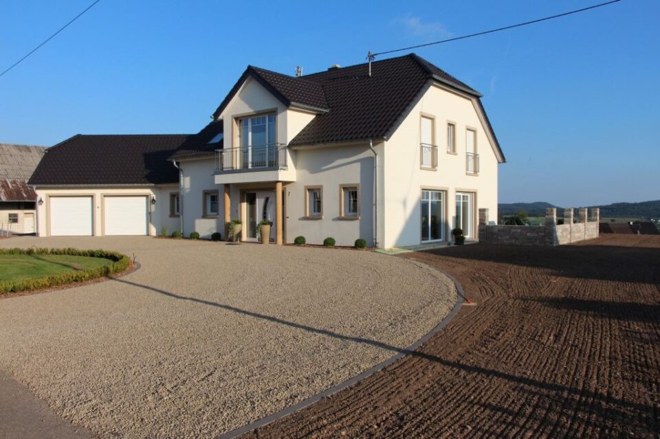 Ein modernes zweistöckiges Burelbach-Haus mit cremefarbenen Wänden, dunklen Dachziegeln, großen Fenstern und einem Balkon. Es verfügt über eine Doppelgarage, eine kreisförmige Kiesauffahrt und ein innovatives Gardendesign in Landschaftsbereichen unter einem klaren blauen Himmel.