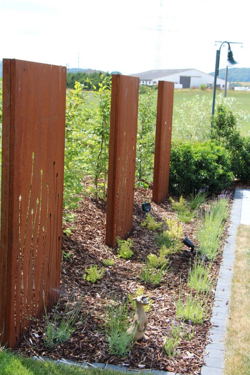 Drei hohe, rostfarbene Metall-Gartenplatten mit ausgeschnittenen Grasmustern stehen zwischen Mulch, Grünpflanzen und einer kleinen Tierstatue - eine künstlerische Note für jede Gartengestaltung - mit einer Wiese und Gebäuden im Hintergrund.