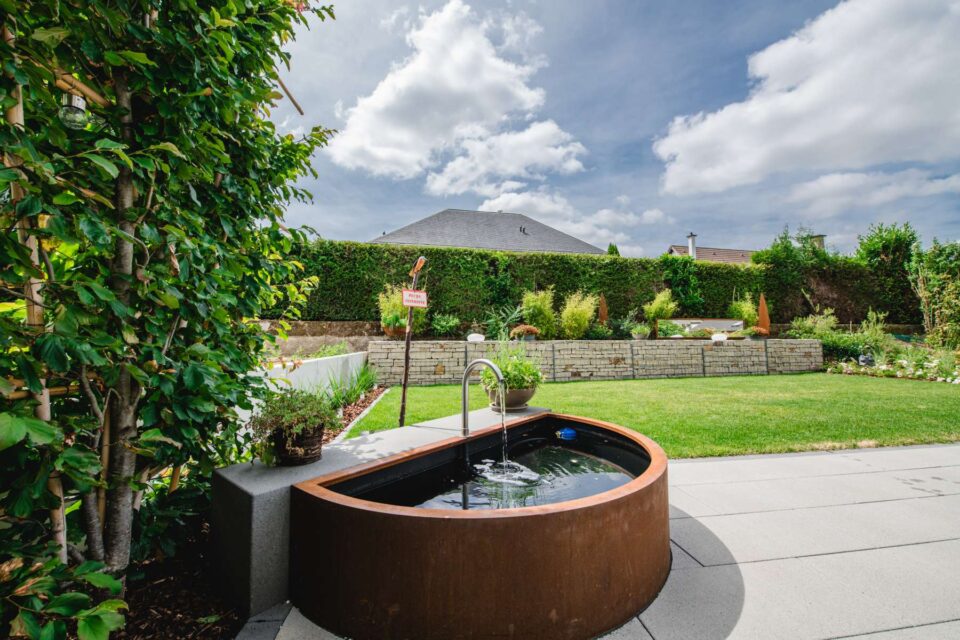 Ein runder, handwerklich gefertigter Whirlpool aus Metall steht auf einer Terrasse mit Blick auf einen grünen Rasen und einen Landschaftsgarten, mit einer Hecke und einem Haus im Hintergrund unter einem teilweise bewölkten Himmel - ganz im Stil von Burelbach in Luxemburg und Deutschland.