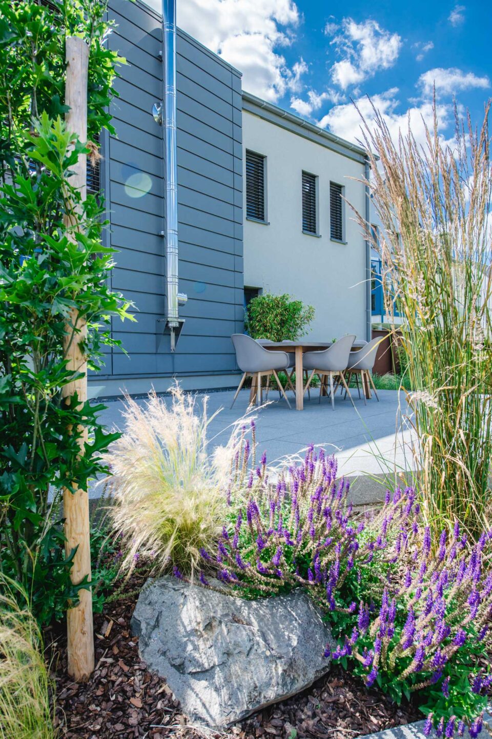 Moderne Terrasse von Burelbach mit grauen Stühlen und einem Holztisch, umgeben von üppigen Grünpflanzen und lila Blumen. Blauer Himmel mit Wolken über einem hellen Haus mit dunklen Fensterläden - perfektes Gartendesign in Luxemburg und Deutschland.