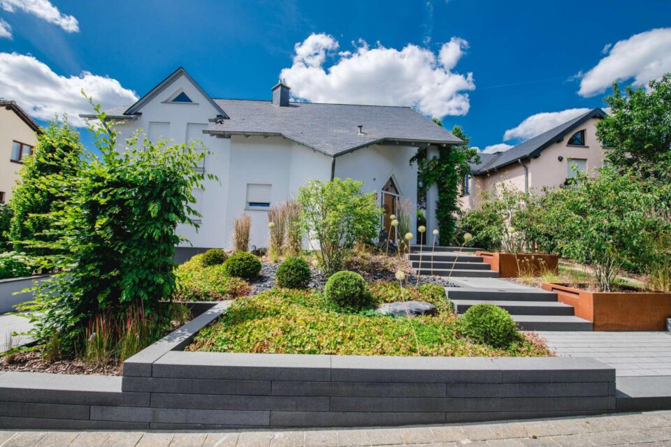 Ein modernes Haus mit grauem Dach und weißen Wänden, umgeben von einem ordentlich angelegten Garten- und Landschaftsbau mit Sträuchern, Büschen und Steinwegen unter einem strahlend blauen Himmel mit vereinzelten Wolken.