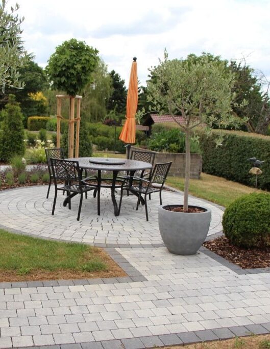 Auf einer runden Steinterrasse stehen Burelbach Gartenmöbel: ein runder Tisch, vier schwarze Metallstühle, ein orangefarbener Sonnenschirm und ein großer Topfbaum, alles in fachmännischer Handarbeit gefertigt und umgeben von üppigem Grün und einem angelegten Garten.