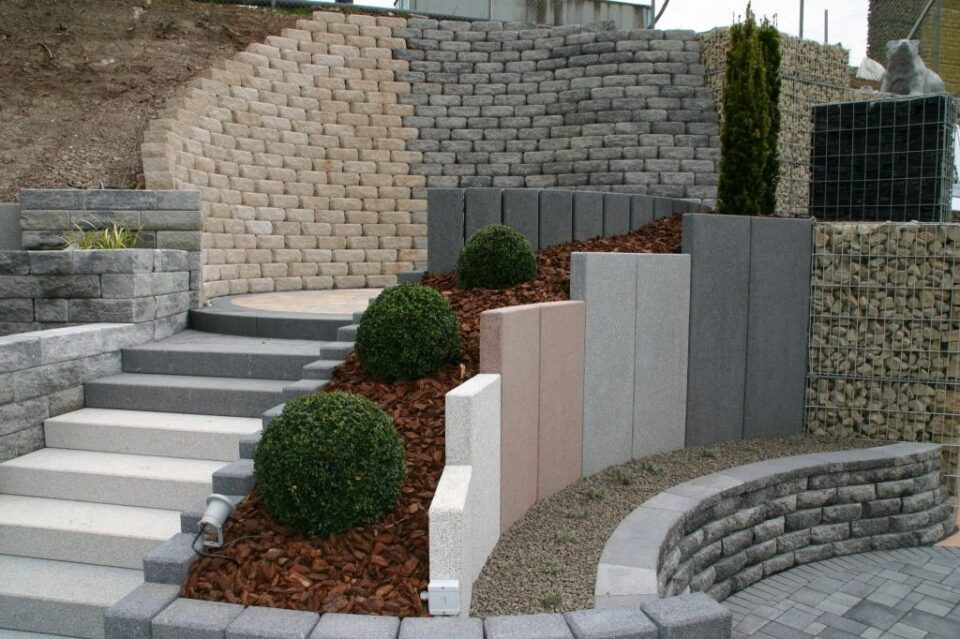 Ein stufenförmiger Garten mit Betonmauern, runden Steinstufen, Kieswegen, vertikalen Dekorationsplatten in neutralen Tönen und ordentlich geschnittenen Rundsträuchern mit braunem Mulch entlang des Hangs - perfekt, um stilvolle Gartenmöbel von Burelbach in Szene zu setzen.