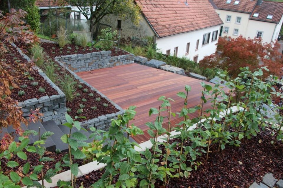 Ein moderner Terrassengarten mit Steinmauern, einem Holzdeck und jungen Grünpflanzen im Vordergrund zeigt fachmännischen Garten- und Landschaftsbau. Im Hintergrund sind Wohnhäuser mit roten Dächern zu sehen.