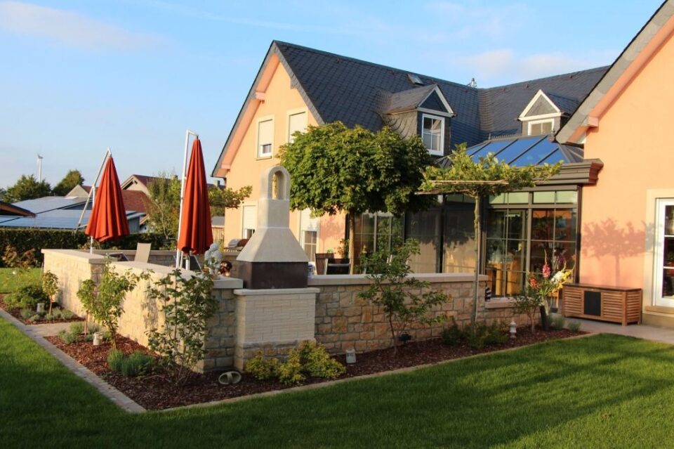 Ein modernes Haus mit Burelbach Gartenmöbeln auf einer Steinterrasse, roten Sonnenschirmen, einem Grill und einem Garten mit Sträuchern und Blumen, alles unter einem blauen Himmel.