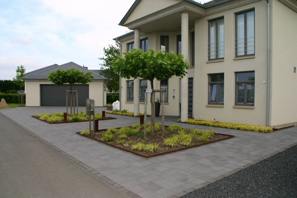 Modernes zweistöckiges Haus mit großen Fenstern, einer gepflasterten Auffahrt, gepflegtem Garten- und Landschaftsbau, kleinen Bäumen vor dem Haus und einer freistehenden Garage auf der linken Seite. Das helle Äußere weist elegante graue Akzente auf.
