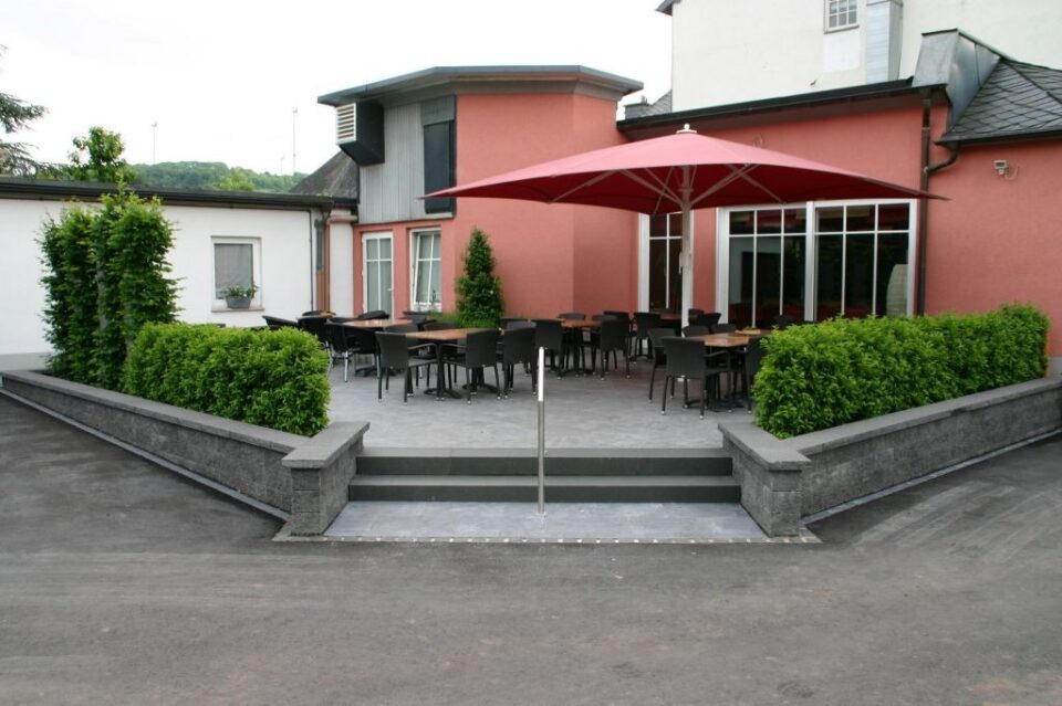 Eine Terrasse mit schwarzen Tischen und Stühlen, rotem Sonnenschirm, umgeben von grünen Hecken und einer niedrigen Steinmauer, die elegantes Gardendesign zeigt, vor einem rosa-weißen Gebäude. Der Bereich ist leer, gut gepflegt, und spiegelt Qualität Handwerk.