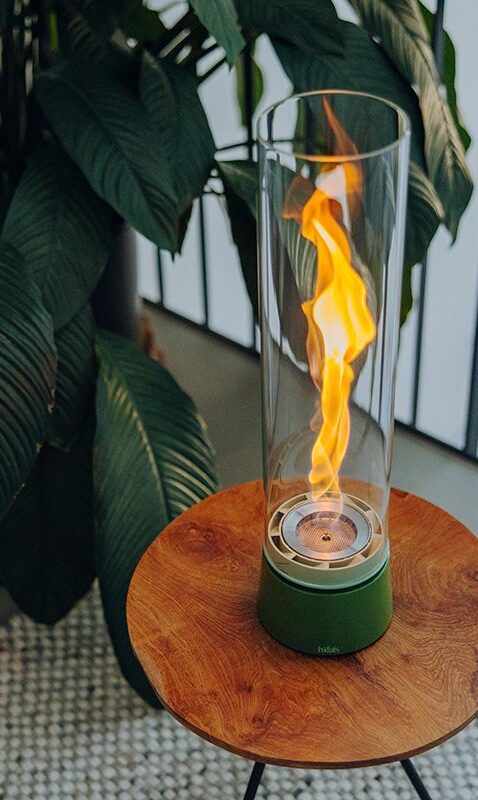 Ein moderner Tischkamin von Burelbach Handwerk mit einem hohen Glaszylinder und sichtbarer Flamme steht auf einem runden Holztisch; große grüne Pflanzenblätter ergänzen das einladende Gardendesign im Hintergrund.
