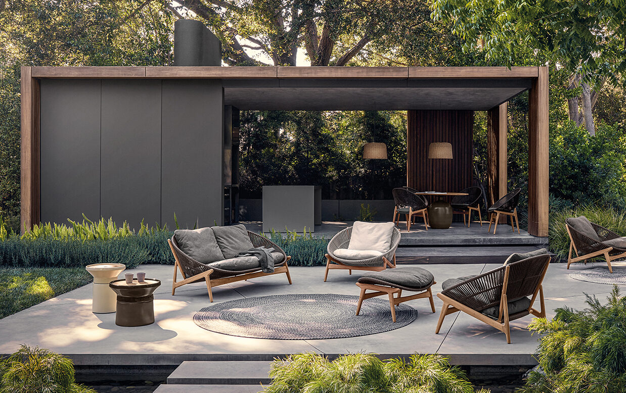 Moderne Außenterrasse mit stilvollen Loungesesseln aus Holz, einem runden Teppich und kleinen Beistelltischen auf einer Betonterrasse. Ein überdachter Essbereich ergänzt das vom Handwerk aus Deutschland und Luxemburg inspirierte Gardendesign, das von üppigem Grün umgeben ist.
