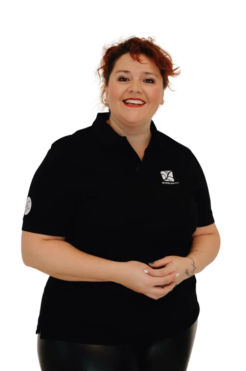 Eine lächelnde Frau mit roten Haaren, die ein schwarzes Poloshirt mit weißem Logo trägt, steht mit gefalteten Händen vor einem weißen Hintergrund und repräsentiert das handwerkliche Können und den Stolz von Garten- und Landschaftsbau und Handwerk.