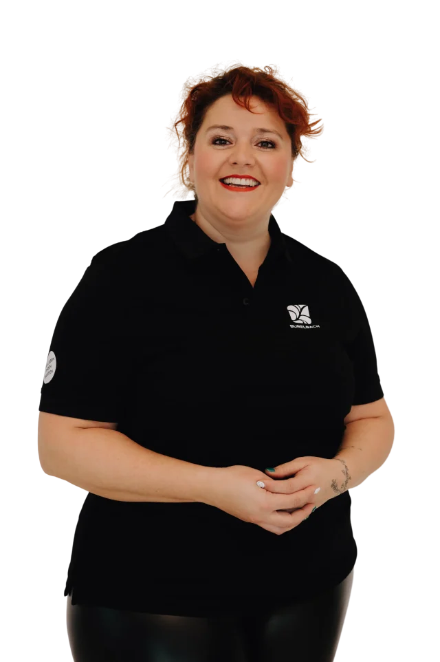 Eine lächelnde rothaarige Frau in einem schwarzen Poloshirt steht mit gefalteten Händen vor einem schlichten Hintergrund und repräsentiert Burelbachs Leidenschaft für Gardendesign und stilvolle Outdoormöbel.