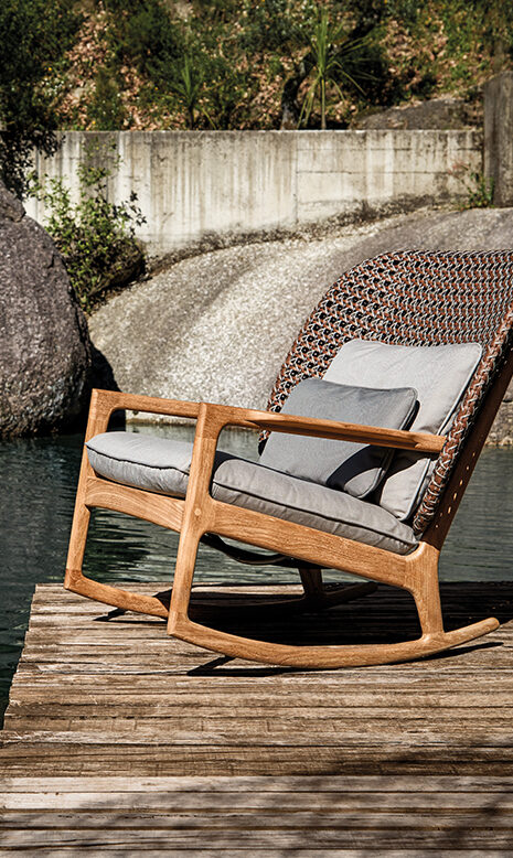 Ein gepolsterter Holzschaukelstuhl mit geflochtener Rückenlehne steht auf einem verwitterten Steg am Wasser, umgeben von Felsen und Grün - die perfekte Kulisse für Gardendesign-Inspiration.