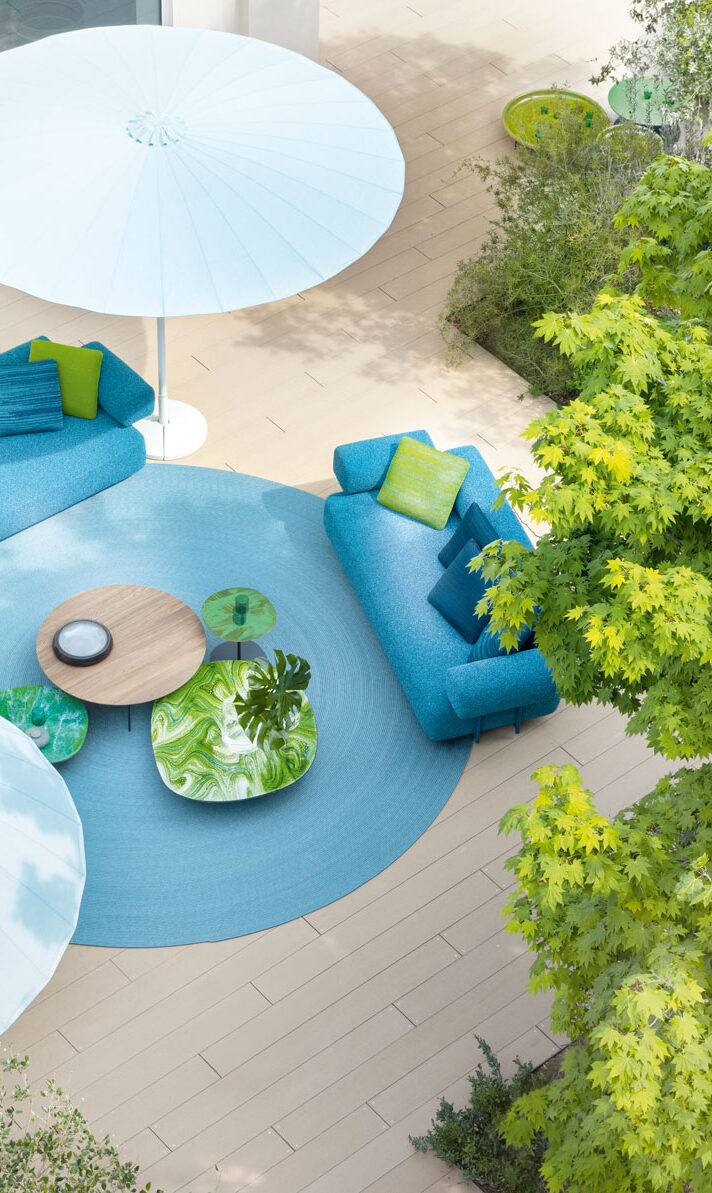 Moderne Außenterrasse mit blauen Sofas, grünen Kissen, rundem Tisch und großen weißen Sonnenschirmen, umgeben von Bäumen. Entdecken Sie stilvolle Outdoormöbel, die Ihr Gardendesign in Deutschland aufwerten.