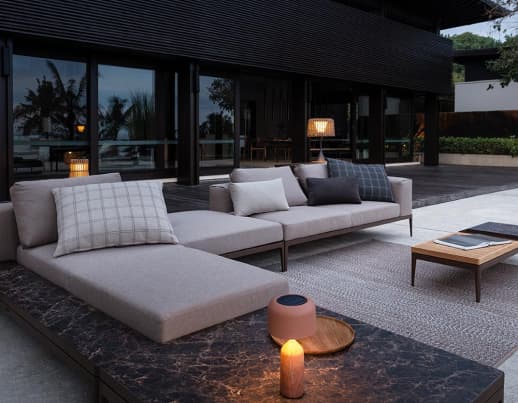 Moderne Außenterrasse von Burelbach mit zwei beigen Lounge-Sofas, neutralen Kissen und einem Marmor-Couchtisch mit Kerzen. Handwerklich inspirierte Details und eine durchdachte Gartengestaltung vervollständigen den Look mit großen Glasfenstern und üppiger Bepflanzung.