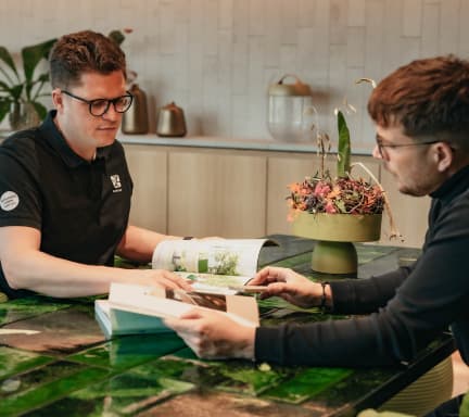 Zwei Männer sitzen an einem Tisch mit grünen Blattmustern und schauen gemeinsam in aufgeschlagene Bücher. Umgeben von Pflanzen und einem Blumenarrangement diskutieren sie über Ideen für die Gartengestaltung. Beide Männer tragen Brillen und dunkle Hemden, was ihre Leidenschaft für den Garten- und Landschaftsbau widerspiegelt.