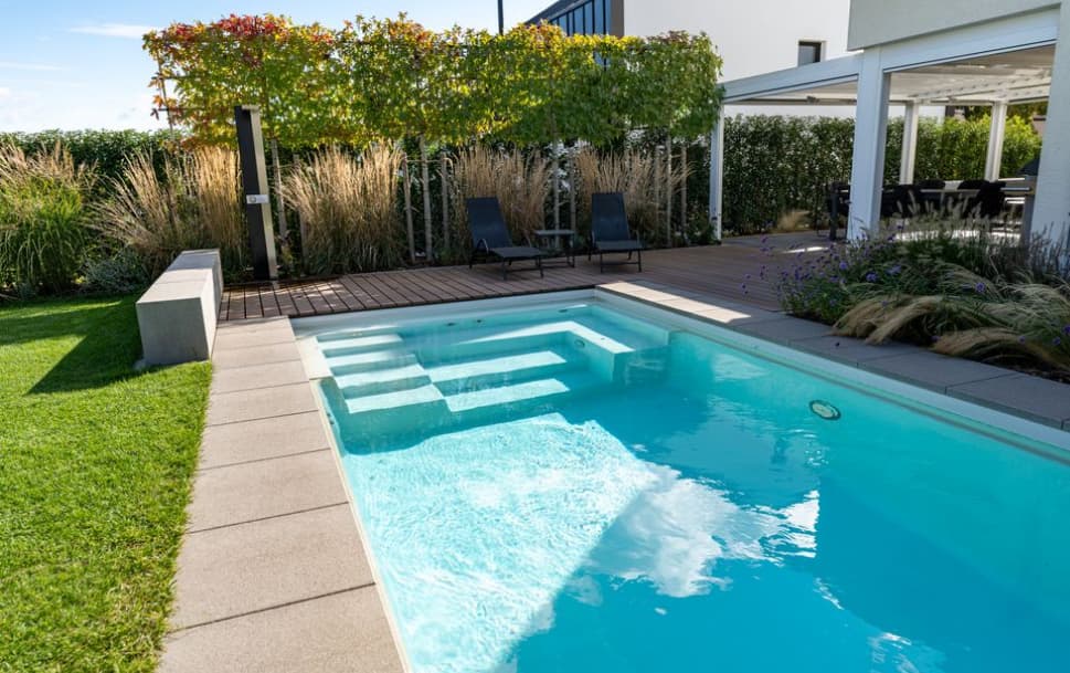 Ein moderner Garten von Burelbach Handwerk verfügt über einen kleinen rechteckigen Swimmingpool mit Stufen, umgeben von Rasen, Terrassen, Liegestühlen und üppigen Pflanzen. Eine überdachte Terrasse mit einem Essbereich vervollständigt diesen eleganten Außenbereich in Deutschland und Luxemburg.