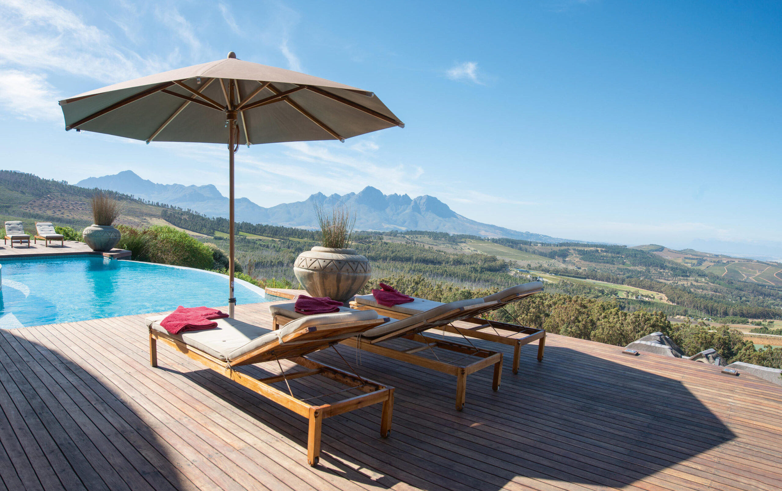 Zwei hölzerne Gartenmöbel mit roten Handtüchern sitzen unter einem großen Sonnenschirm neben einem Infinity-Pool, mit Blick auf sanfte Hügel und ferne Berge - eine atemberaubende Außenszene von Burelbach Garten- und Landschaftsbau an einem klaren, sonnigen Tag.