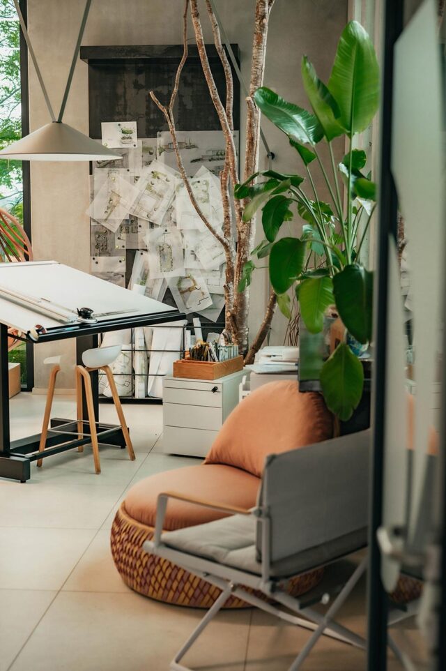 Ein gemütlicher, moderner Arbeitsbereich mit Pflanzen, einem Zeichentisch, Skizzen an der Wand und einem gepolsterten Stuhl mit stilvollen Outdoormöbeln, inspiriert von Trends aus Deutschland und Luxemburg.