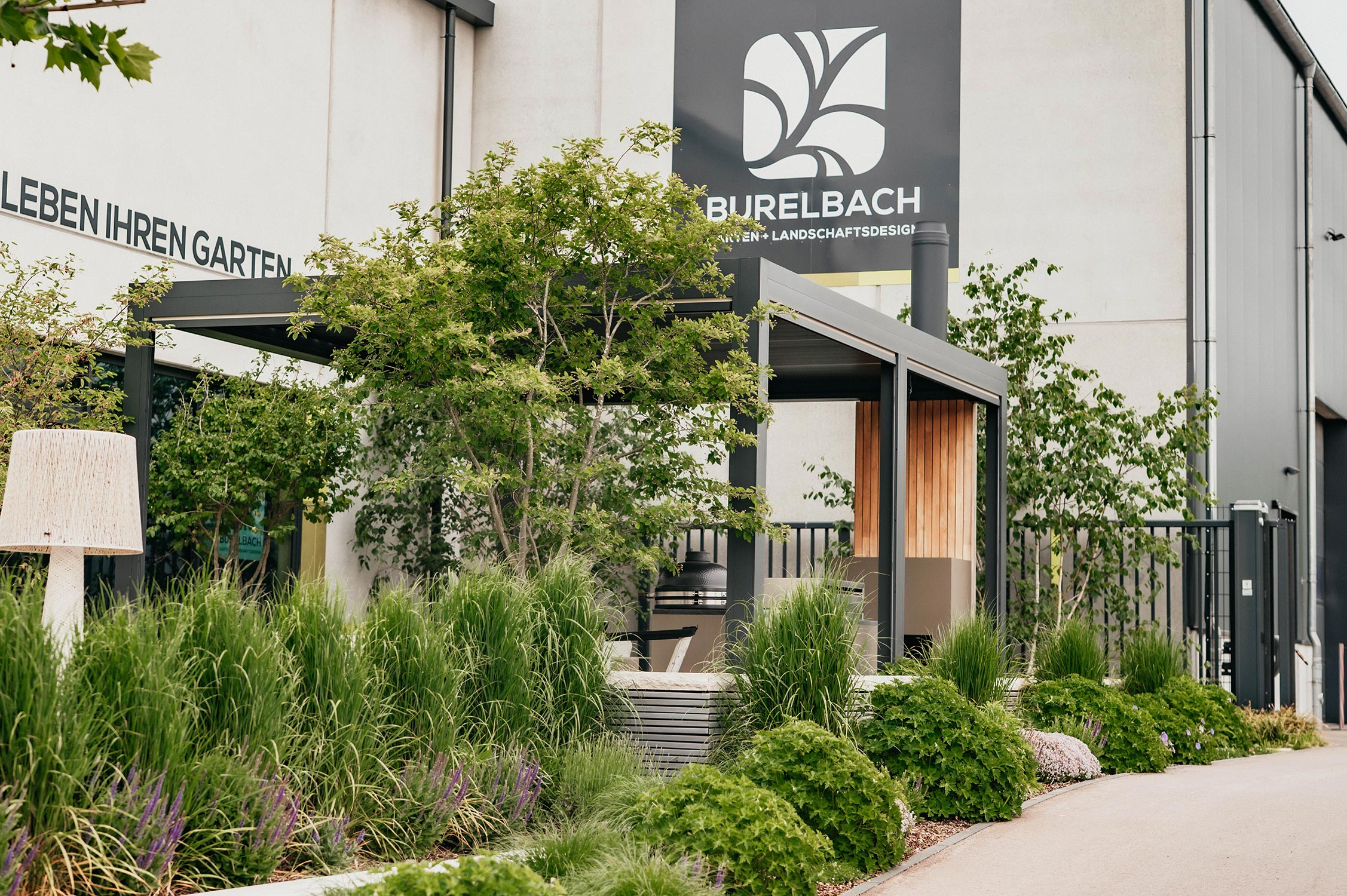 Moderne Gartengestaltung mit üppigen Grünpflanzen, hohen Gräsern und einem schattigen Sitzbereich vor einem Gebäude mit einem "Burelbach"-Schild. Die Umgebung wirkt sauber und gepflegt.