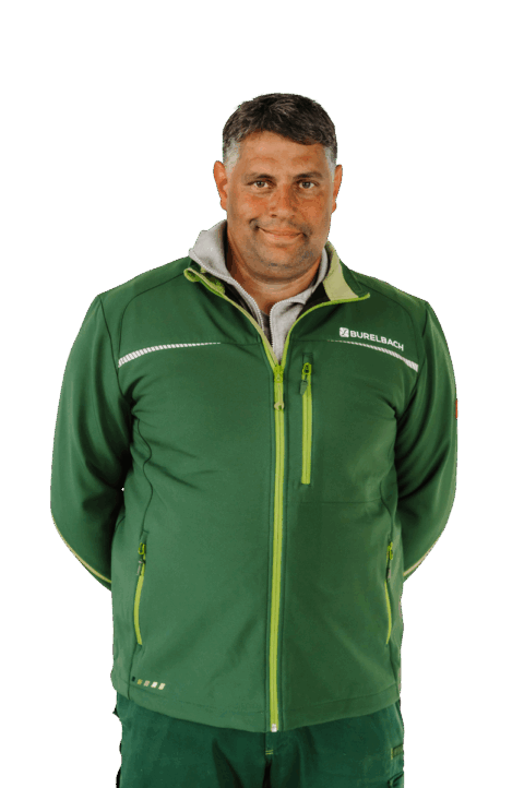 Ein Mann mit kurzen dunklen Haaren, bekleidet mit einer grünen Reißverschlussjacke mit Burelbach-Logo steht vor der Kamera und lächelt leicht, die Hände hinter dem Rücken.