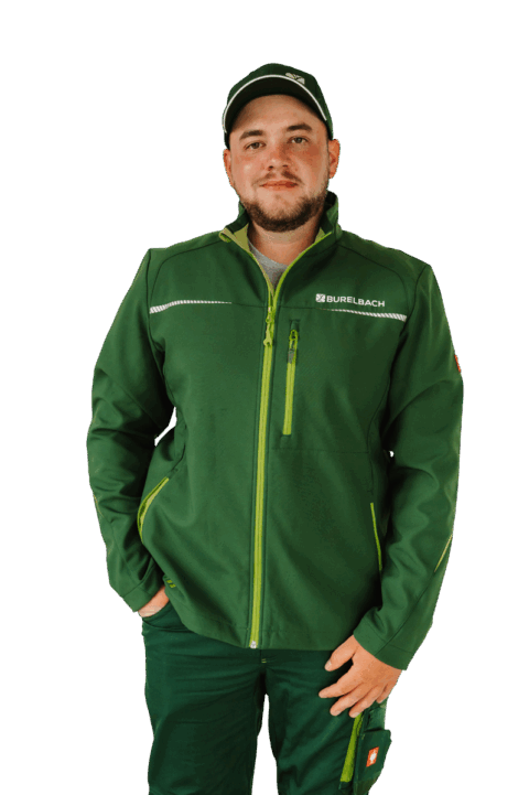 Ein Mann mit grüner Mütze und grüner Arbeitsjacke mit dem "BURELBACH"-Logo steht mit dem Gesicht nach vorne, eine Hand in der Tasche, und schaut mit neutralem Blick in die Kamera.
