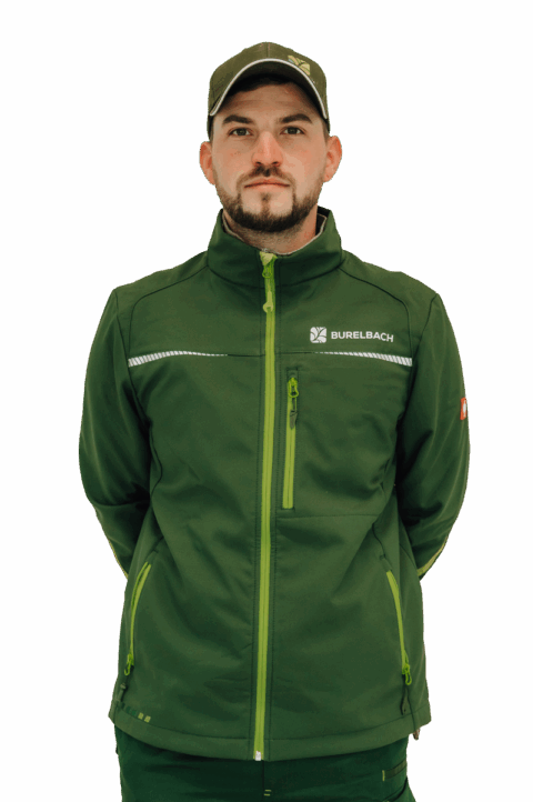 Ein Mann mit grüner Mütze und grüner Jacke mit Reißverschluss und BURELBACH-Logo steht mit dem Gesicht nach vorne und den Händen auf dem Rücken. Der Hintergrund ist einfarbig und weiß.