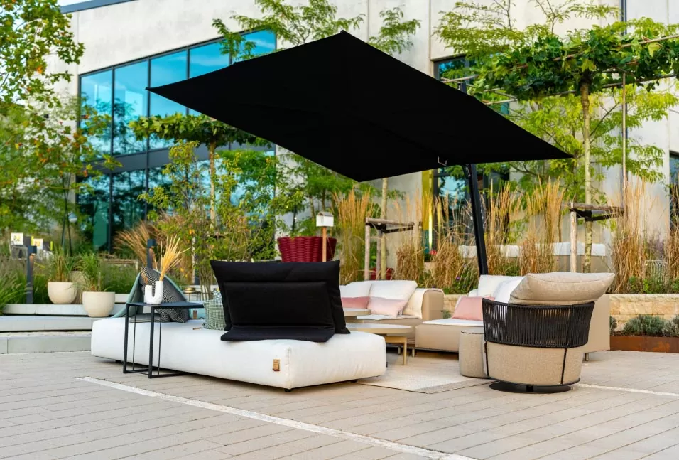 Moderner Outdoor-Loungebereich mit Sofas, Stühlen und einem großen schwarzen Schirm auf einer Terrasse in der Nähe eines Gebäudes, der elegantes Gardendesign von Burelbach in Luxemburg zeigt.