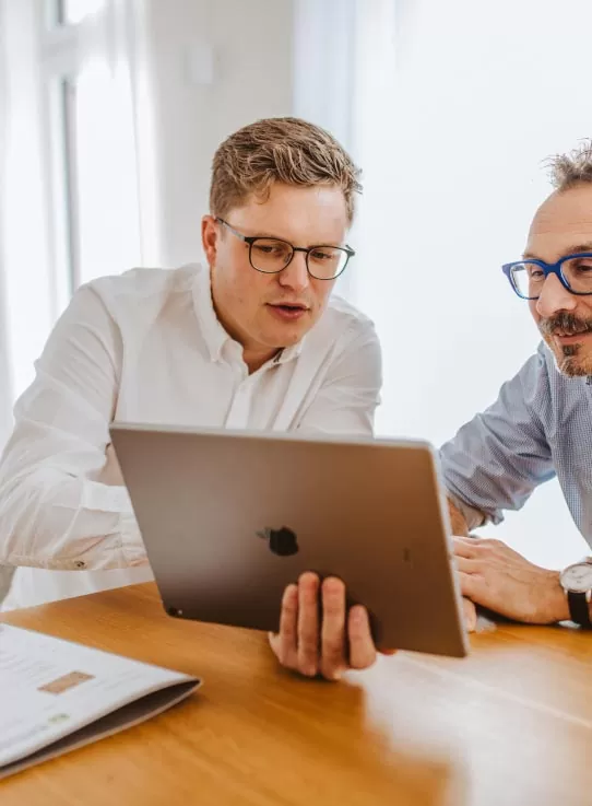 Zwei Männer mit Brillen sitzen an einem Tisch und schauen gemeinsam auf einen Laptop in einem hellen Raum mit stilvollen Outdoormöbeln und modernen Gardendesign-Elementen.