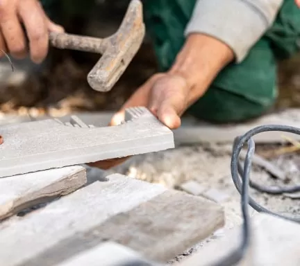 Eine Person benutzt einen Hammer, um ein Stück Stein oder eine Fliese zu formen oder zu zerbrechen - eine Szene, die von handwerklichem Geschick zeugt - während andere Fliesen und Werkzeuge auf der Arbeitsfläche im Freien liegen, was auf Garten- und Landschaftsbauaktivitäten hindeutet.