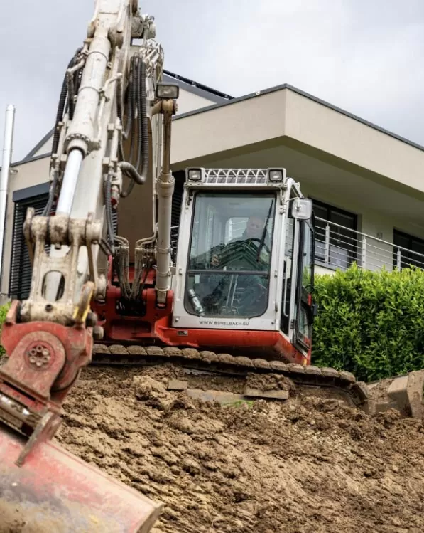 Ein rot-weißer Bagger gräbt Erde vor einem modernen Haus mit großen Fenstern und einem Balkon, umgeben von Grün - eine beeindruckende Szene, die Handwerk in Luxemburg und Deutschland unter einem bewölkten Himmel zeigt.