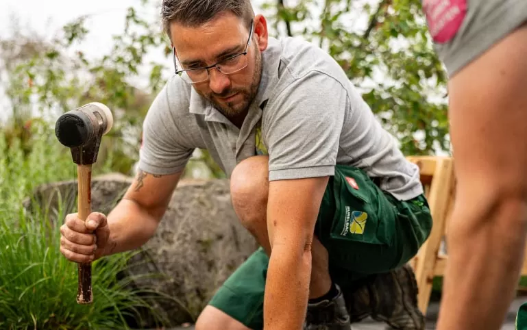 Ein Mann mit Brille und Arbeitskleidung kniet im Freien, hält einen Gummihammer und bearbeitet den Boden. Im Hintergrund sind Grünpflanzen und felsige Landschaftsgestaltung von Burelbach Garten- und Landschaftsbau zu sehen.