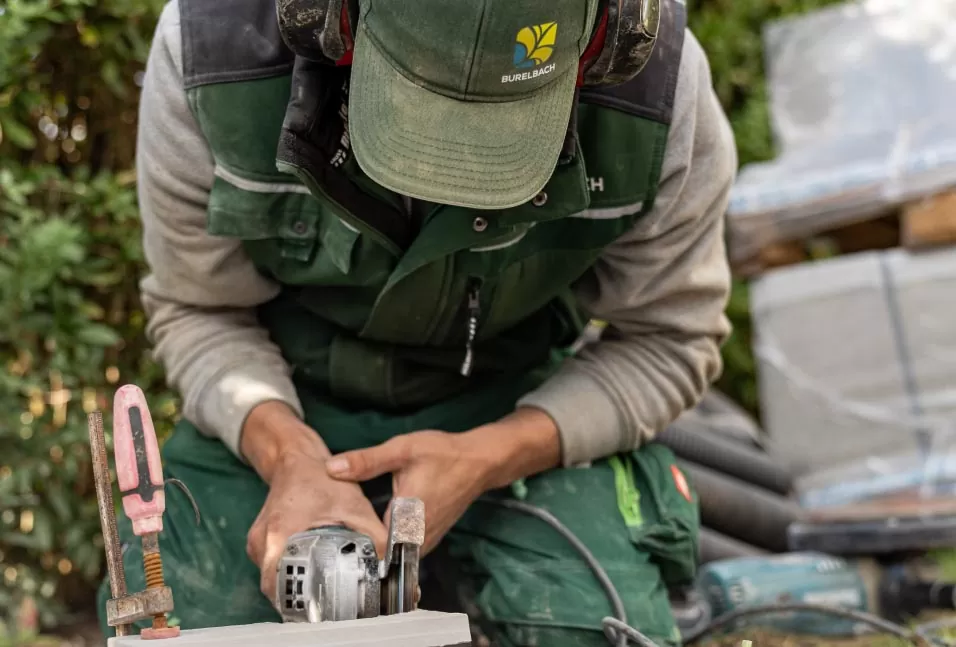 Ein Arbeiter in grüner Uniform schneidet mit einem handgeführten Elektrowerkzeug eine Steinplatte im Freien und zeigt damit sein handwerkliches Können. Er trägt Gehörschutz und andere Sicherheitsausrüstung. Im Hintergrund sind Grünpflanzen und mögliche Gardendesign-Elemente zu sehen.
