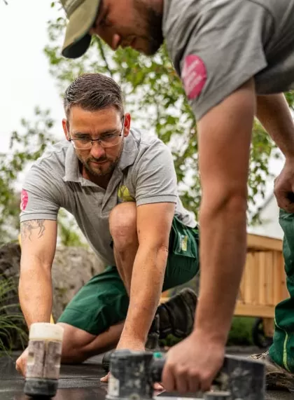 Zwei Männer in grauen Hemden und grünen Shorts knien im Freien und arbeiten eng an einer Fläche mit Werkzeugen. Konzentriert auf ihr Handwerk, trägt einer eine Brille. Bäume und eine Holzkonstruktion für Gartenmöbel sind unscharf im Hintergrund.