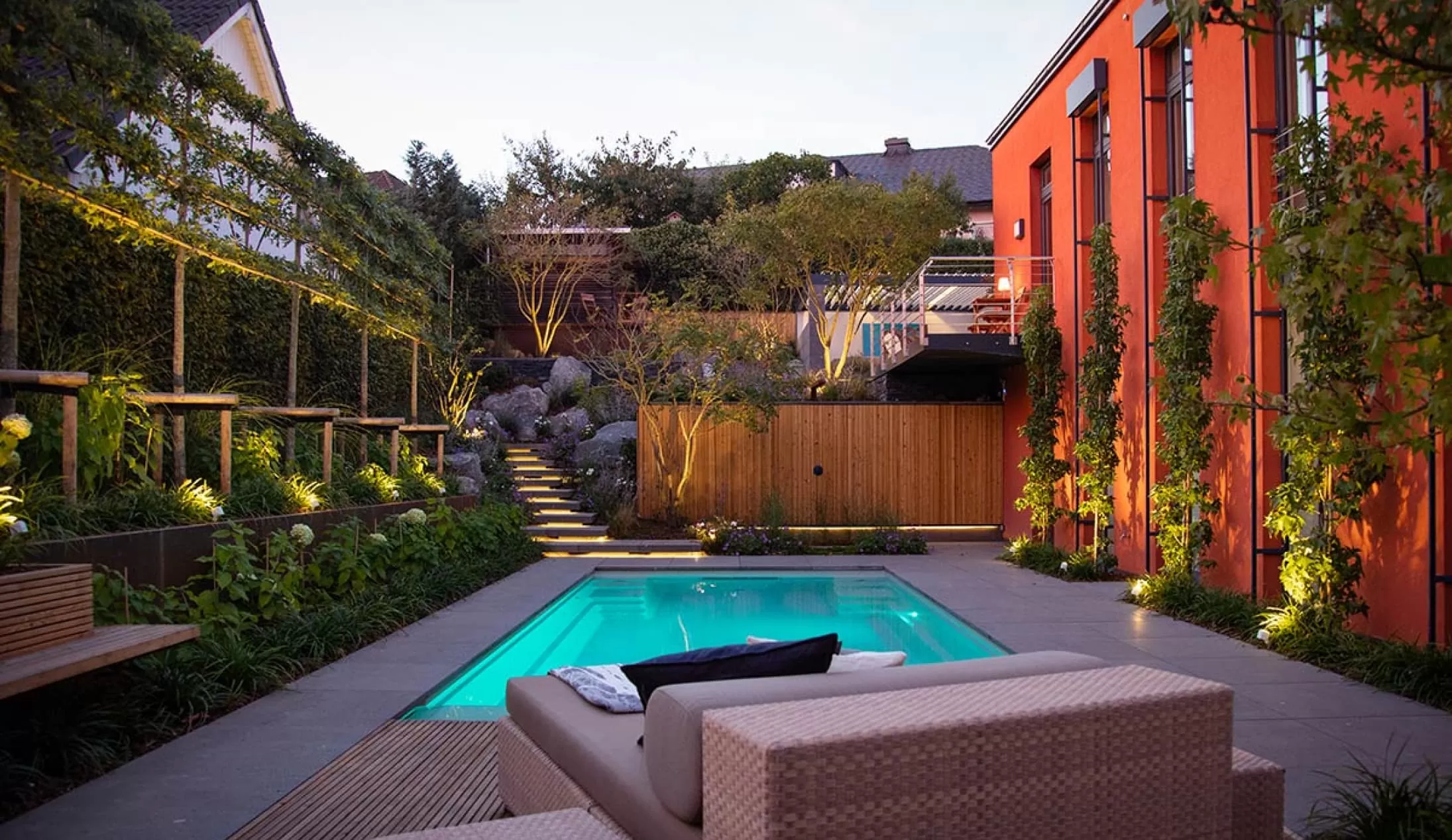 Ein moderner Garten mit rechteckigem Pool von Garten- und Landschaftsbau Profis aus Luxemburg und Deutschland mit üppigem Grün, einem beigen Lounge-Sessel und einer warmen Wegbeleuchtung, die in der Abenddämmerung eine gemütliche, einladende Atmosphäre schafft.
