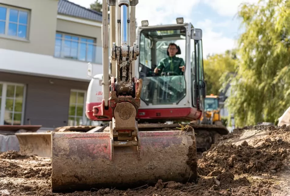 Person, die einen Bagger bedient und auf einer Baustelle vor einem Gebäude Erde aushebt, um Gartenbauprojekte in Deutschland zu unterstützen.