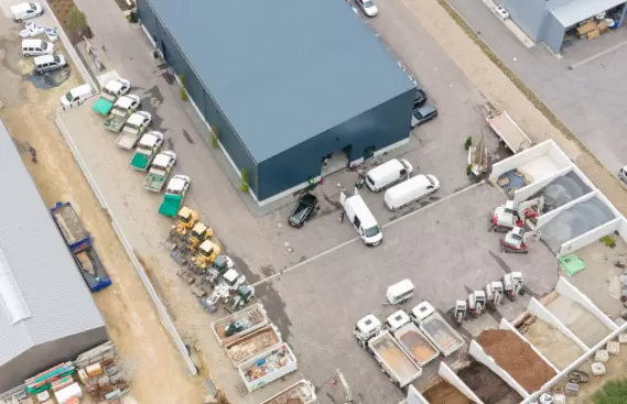 Luftaufnahme einer Industrieanlage in Deutschland mit Lastwagen, Lieferwagen, Lagerplätzen für Baumaterialien und Flächen für Gartenbau und Outdoormöbel.