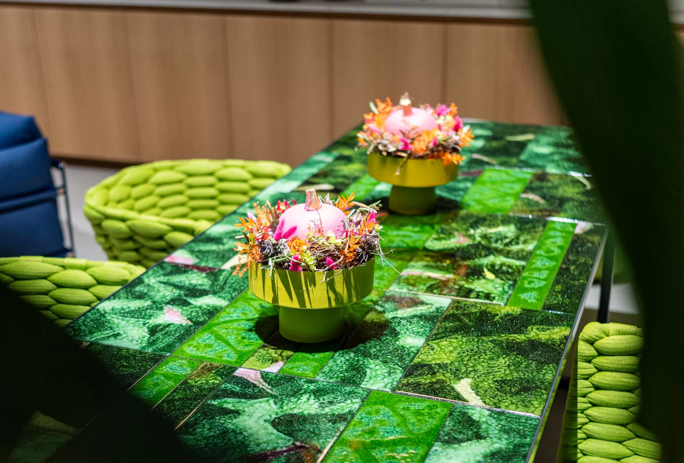 Ein grüner Gartenmöbeltisch mit Blättermuster und zwei passenden Stühlen. Auf dem Tisch stehen farbenfrohe Blumensträuße in grünen Töpfen mit je einer rosafarbenen Dekokugel und bunten Kunstblumen - ideal für Burelbach-Häuser in Luxemburg und Deutschland.