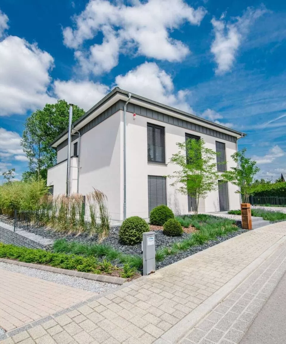 Modernes weißes Haus mit großen Fenstern, stilvollen Outdoormöbeln, einem von Gartenbauexperten angelegten Garten und einem blauen Himmel mit vereinzelten Wolken darüber.